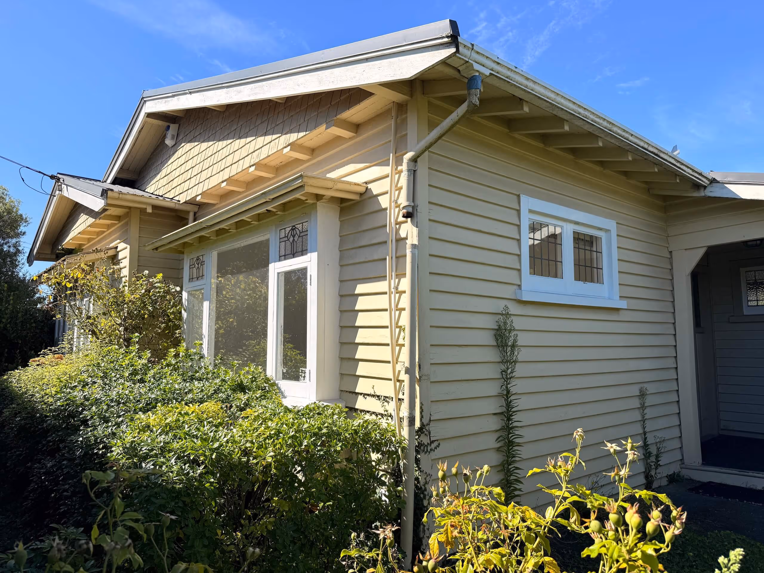 Yellow wooden house with white trim, decorative windows, and green shrubs in front under a clear blue sky.