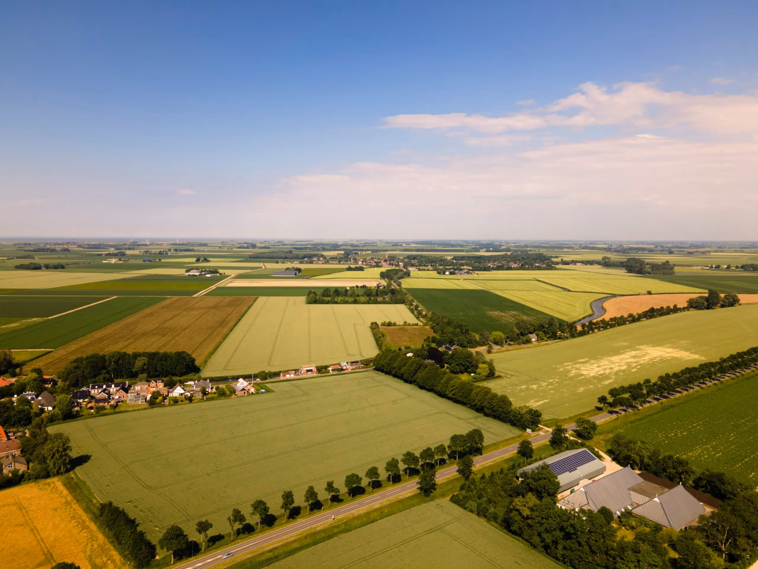 Aerial view of rural farmland with green and yellow fields, scattered houses, tree-lined roads, and a partly cloudy blue sky.