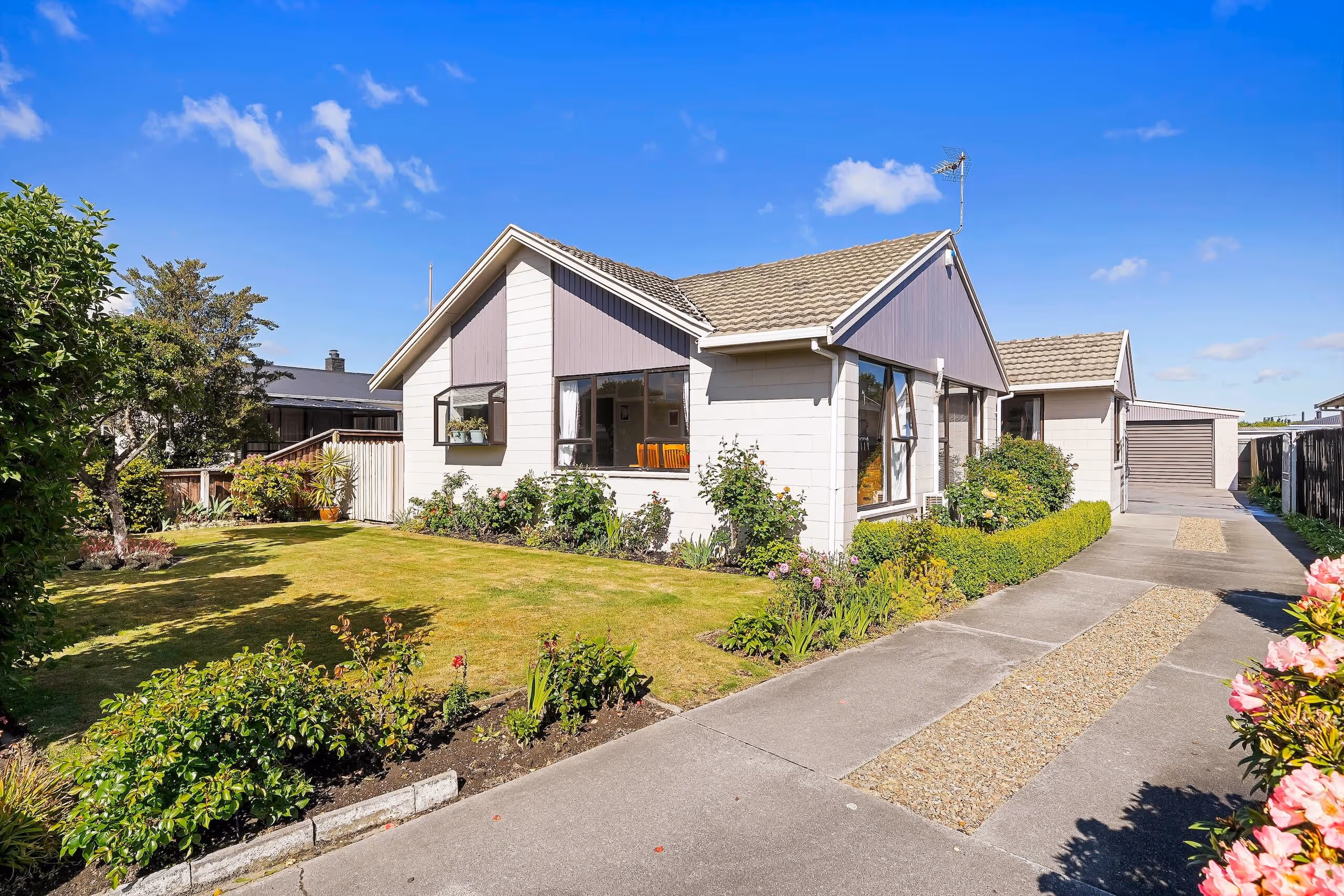 Single-story suburban house with a tiled roof, large windows, front garden with shrubs and flowers, and a driveway leading to a detached garage under a clear blue sky.