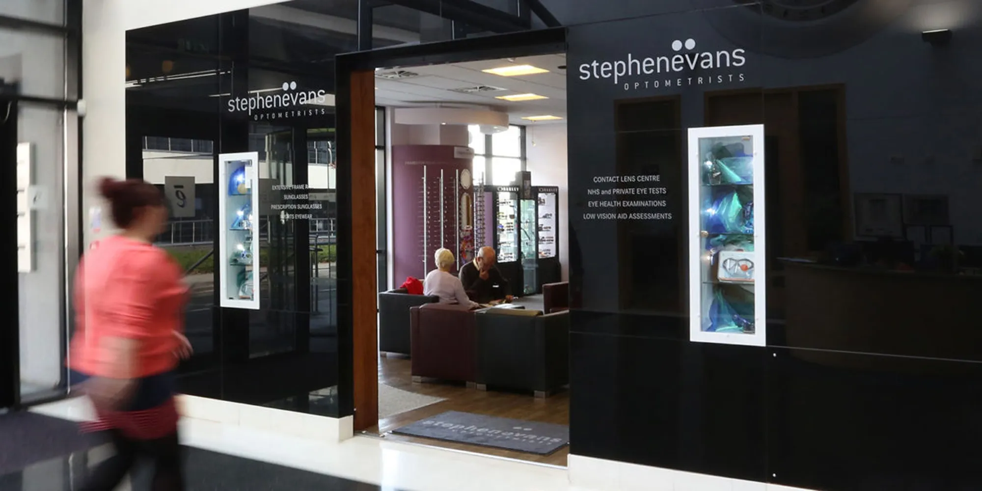 Inside an optometrist office, two people sit discussing at a table while a person walks by outside.