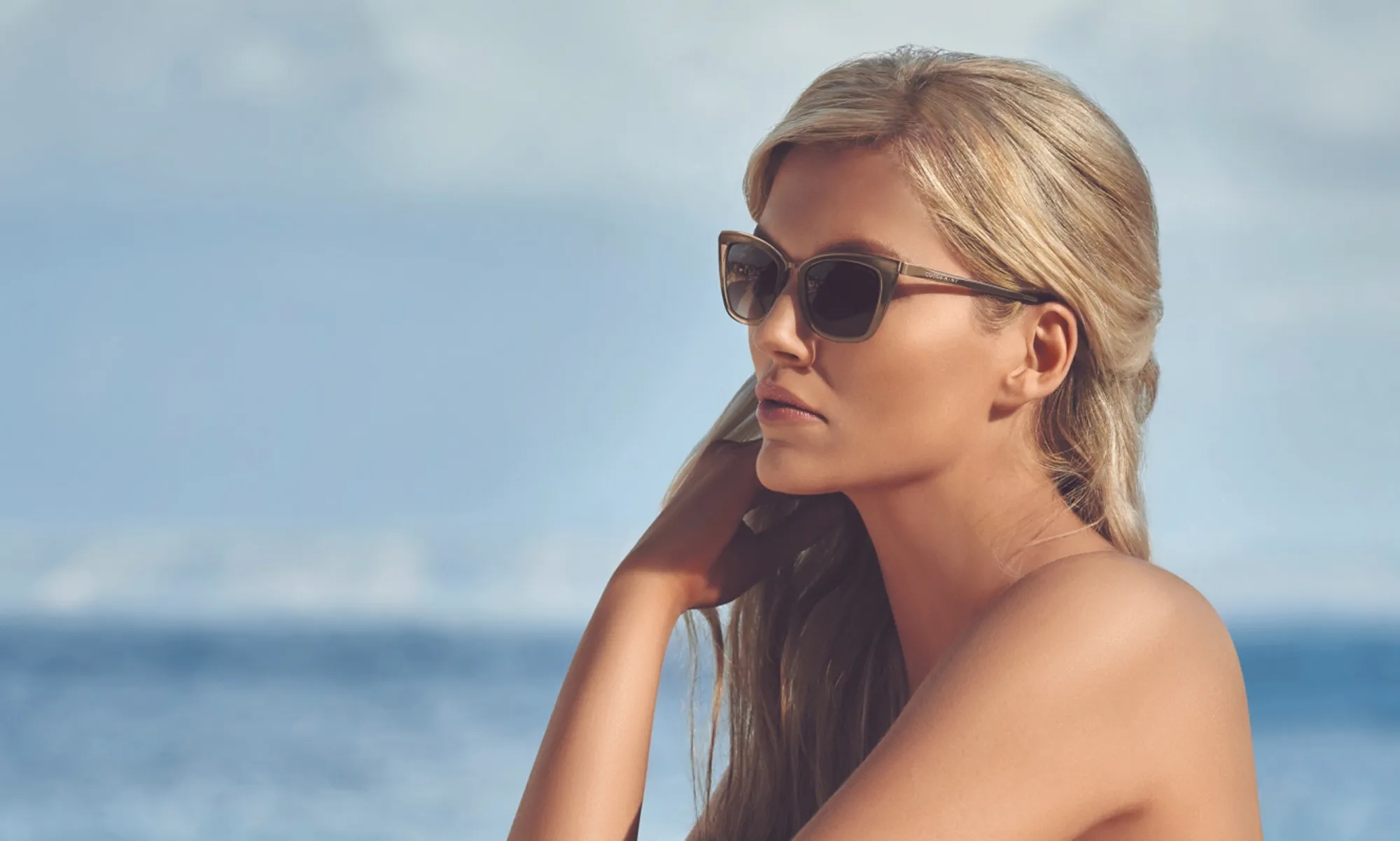 Blonde woman wearing cat-eye sunglasses in profile against a blurred ocean and sky background