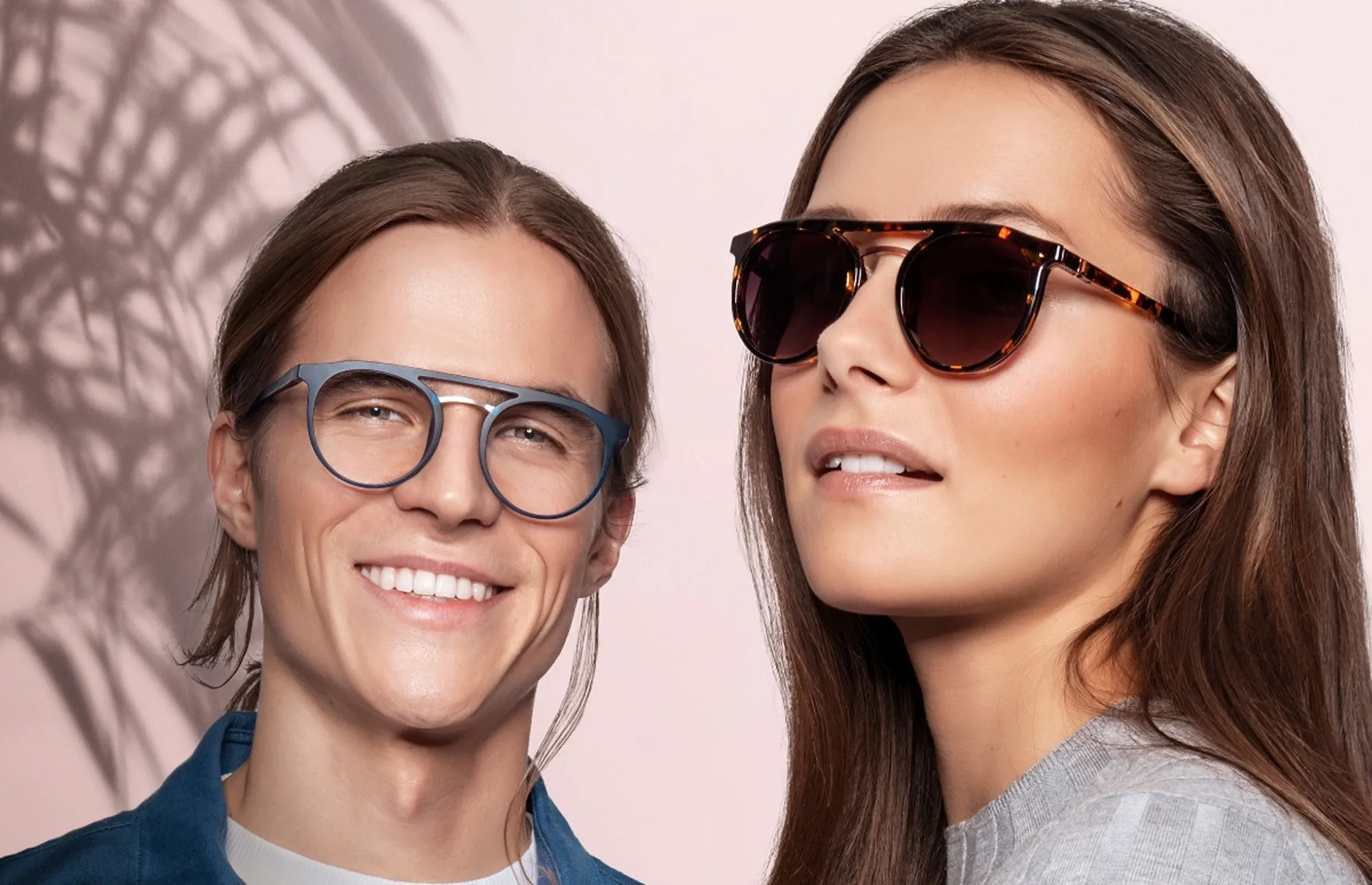 Man in blue eyeglasses and woman in tortoiseshell sunglasses against a pink background with palm shadows.