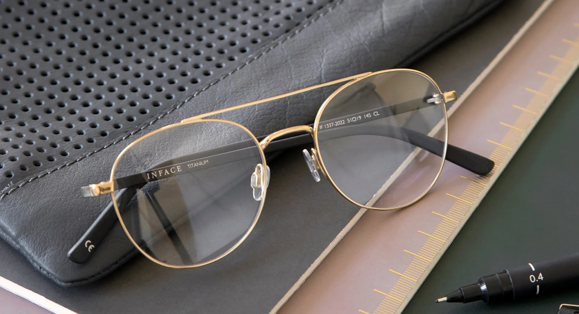 Gold titanium Inface aviator eyeglasses resting on a leather case beside a ruler and pen.