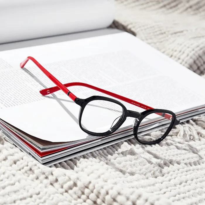 Black eyeglasses with red temples resting on an open magazine and a knit blanket.