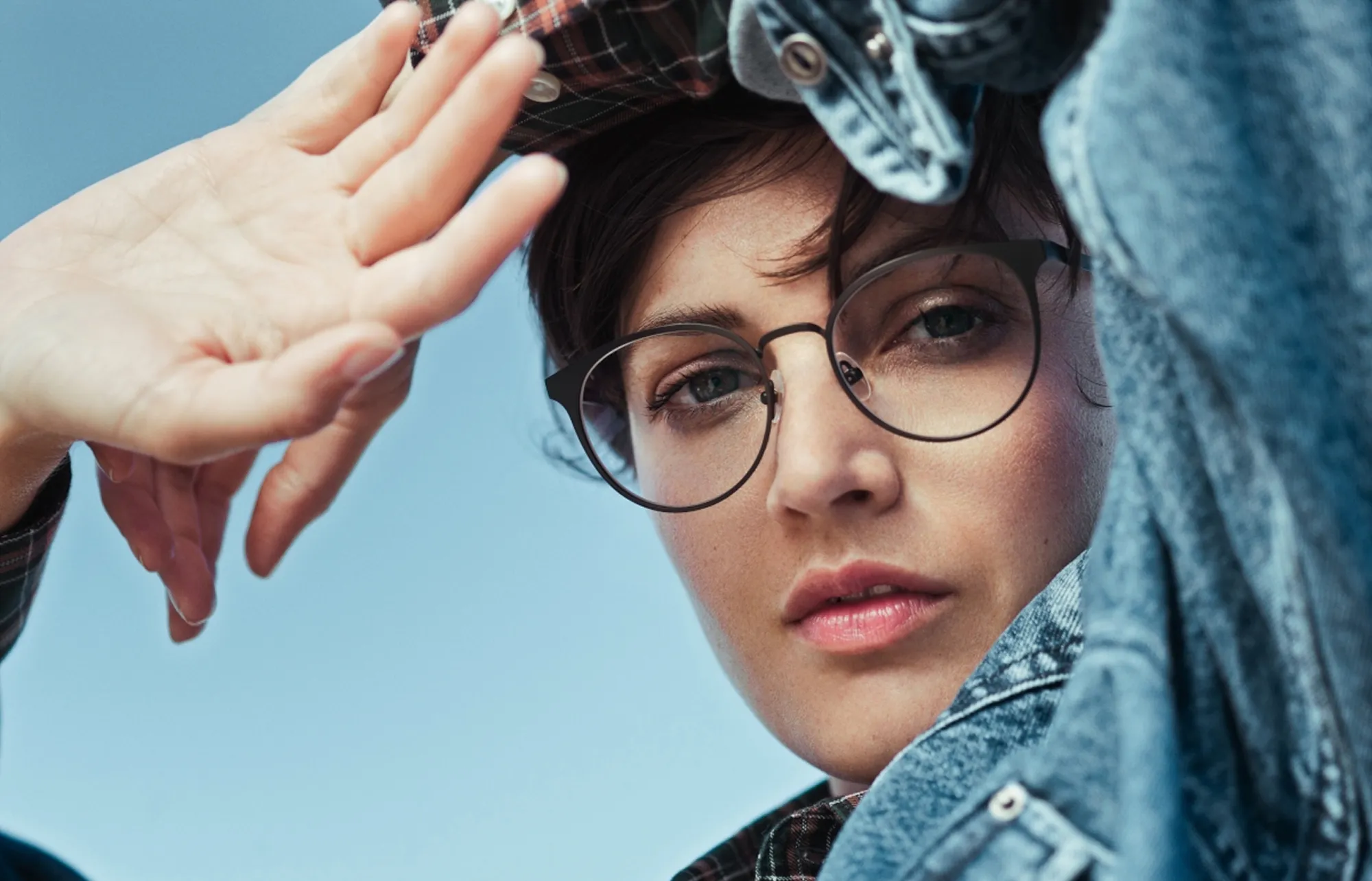 Close-up of a person in round black eyeglasses and a denim jacket against a blue background.