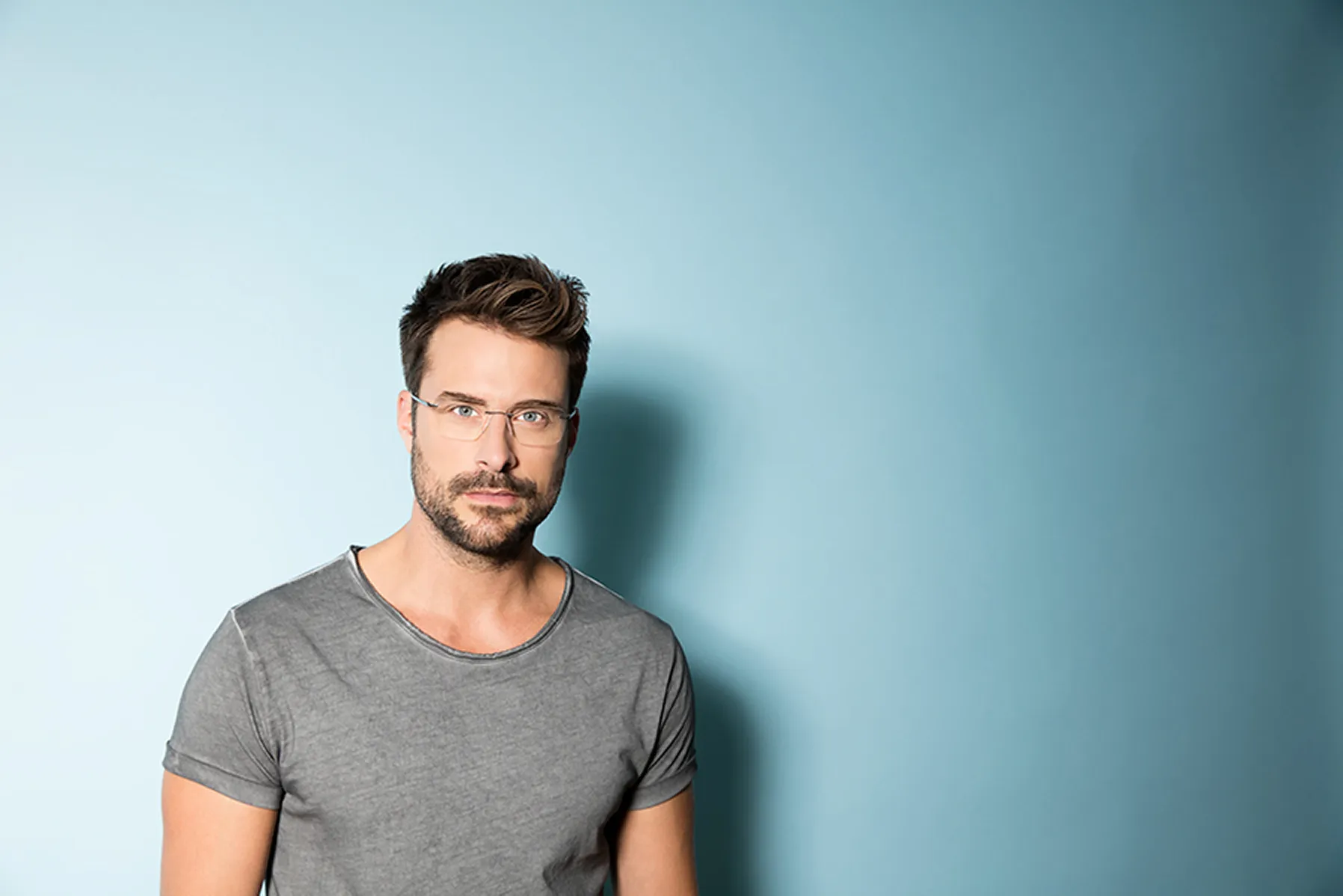 Man in thin rimless eyeglasses and a gray t-shirt against a blue background.