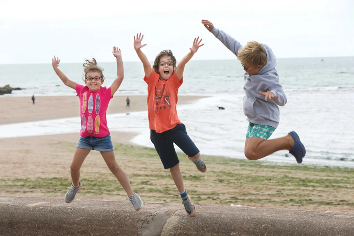 Three children in glasses jumping joyfully on a beach with the sea behind them.