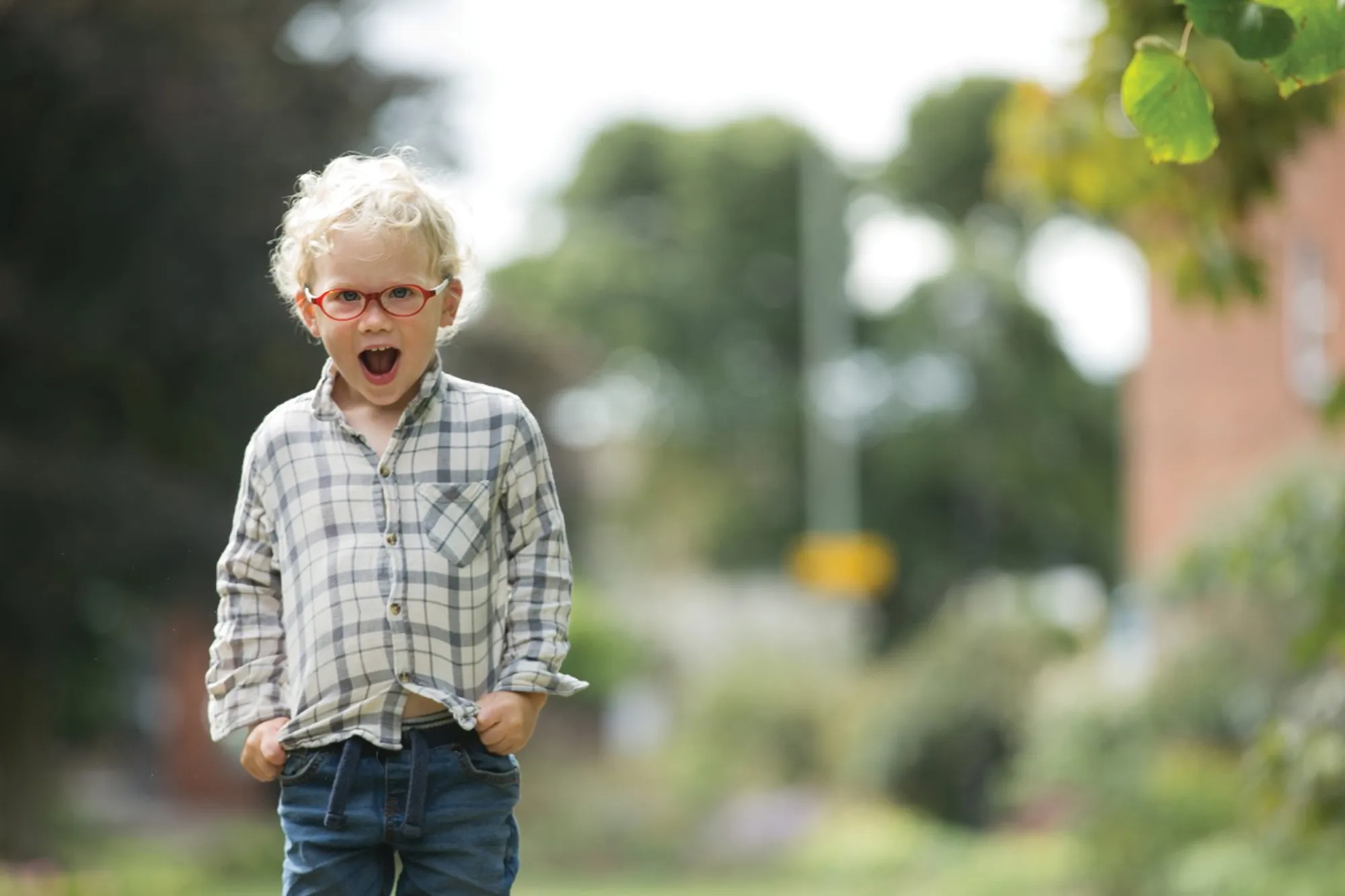 Young child in red glasses and a plaid shirt standing outdoors with a surprised expression.