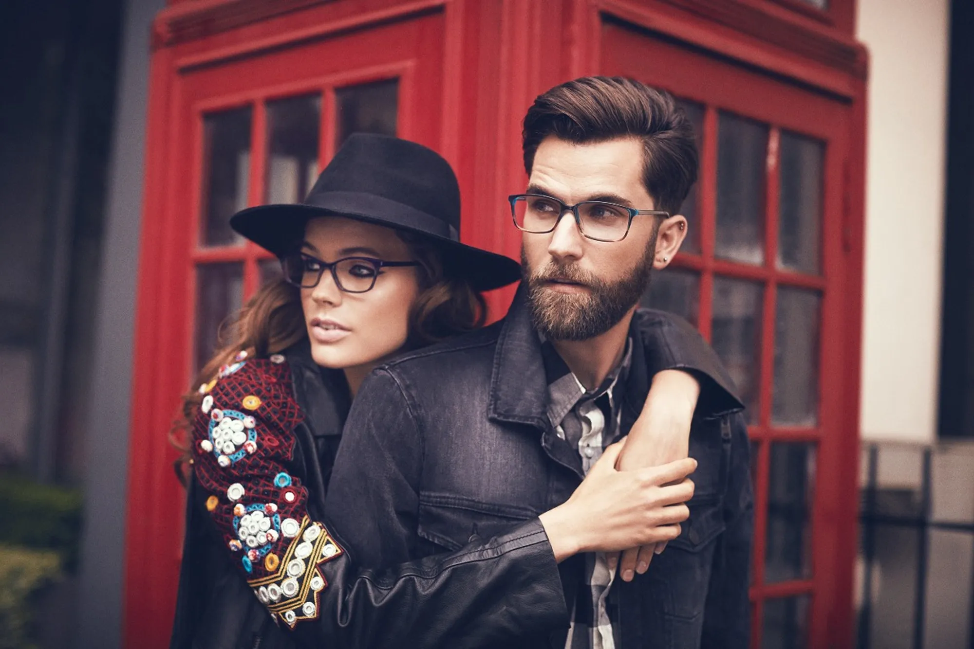 Stylish couple wearing eyeglasses in front of a red telephone booth.
