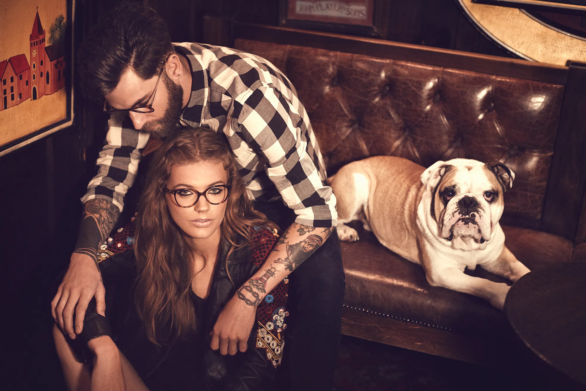Couple wearing eyeglasses beside a bulldog on a leather sofa in a dimly lit interior.