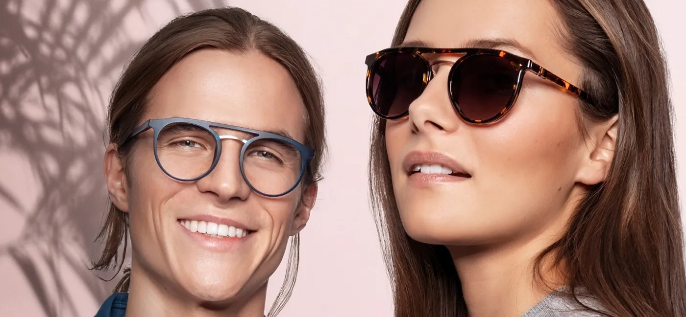 Man wearing blue eyeglasses and woman wearing tortoiseshell sunglasses against a pink background