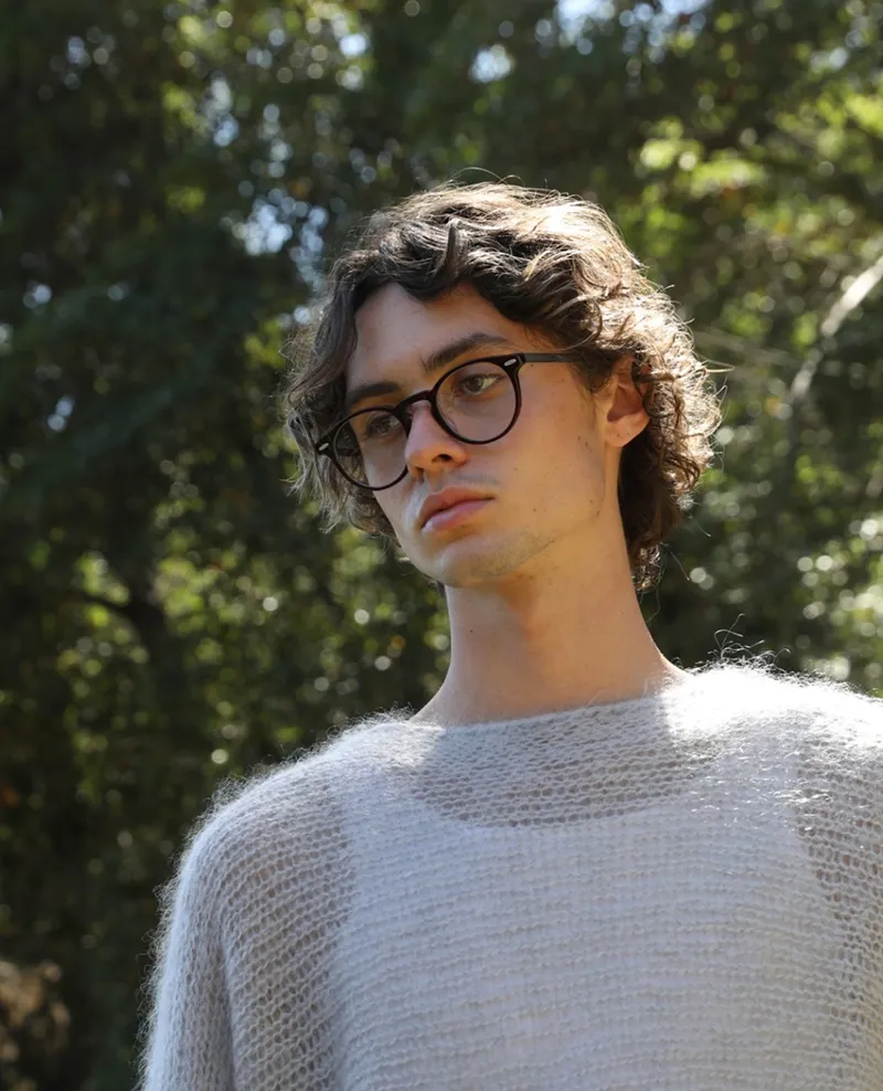 Man with curly hair wearing round black eyeglasses outdoors against greenery