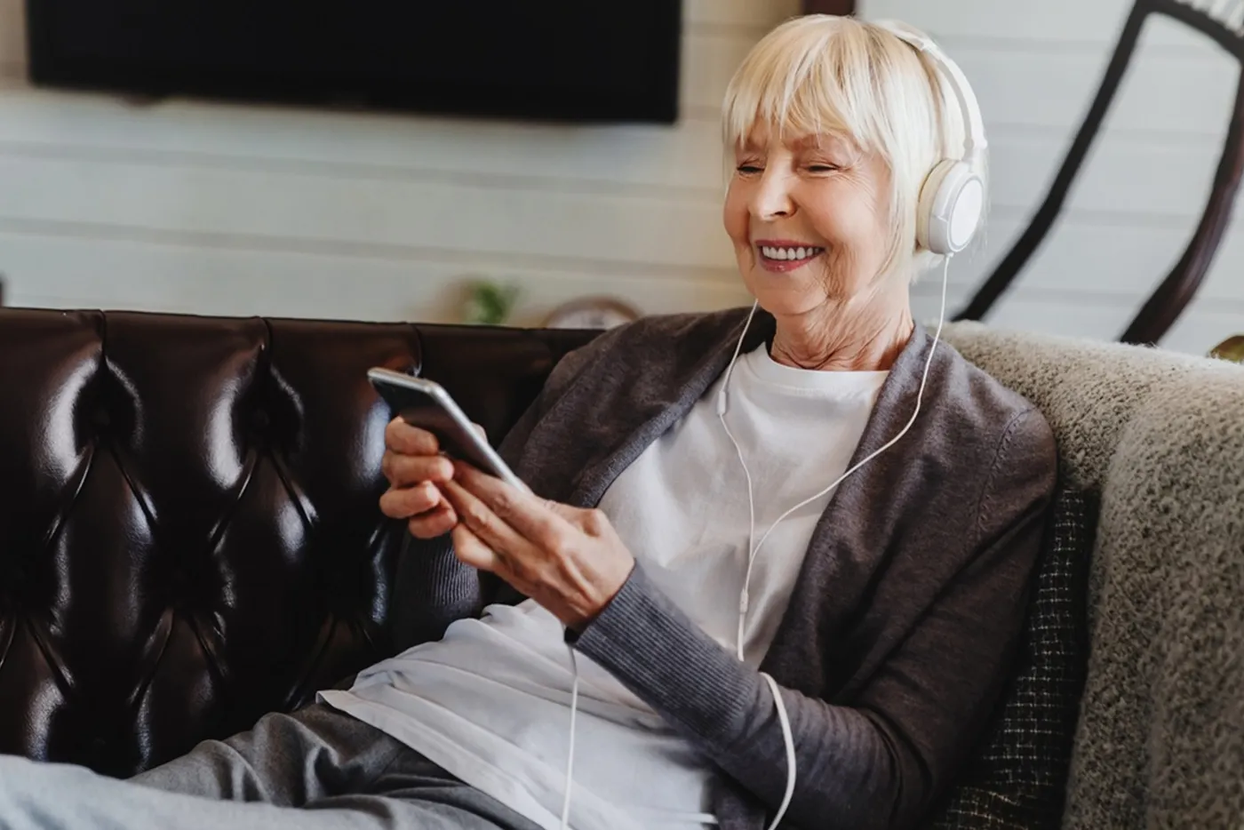 Older woman wearing headphones and smiling at her smartphone on a sofa
