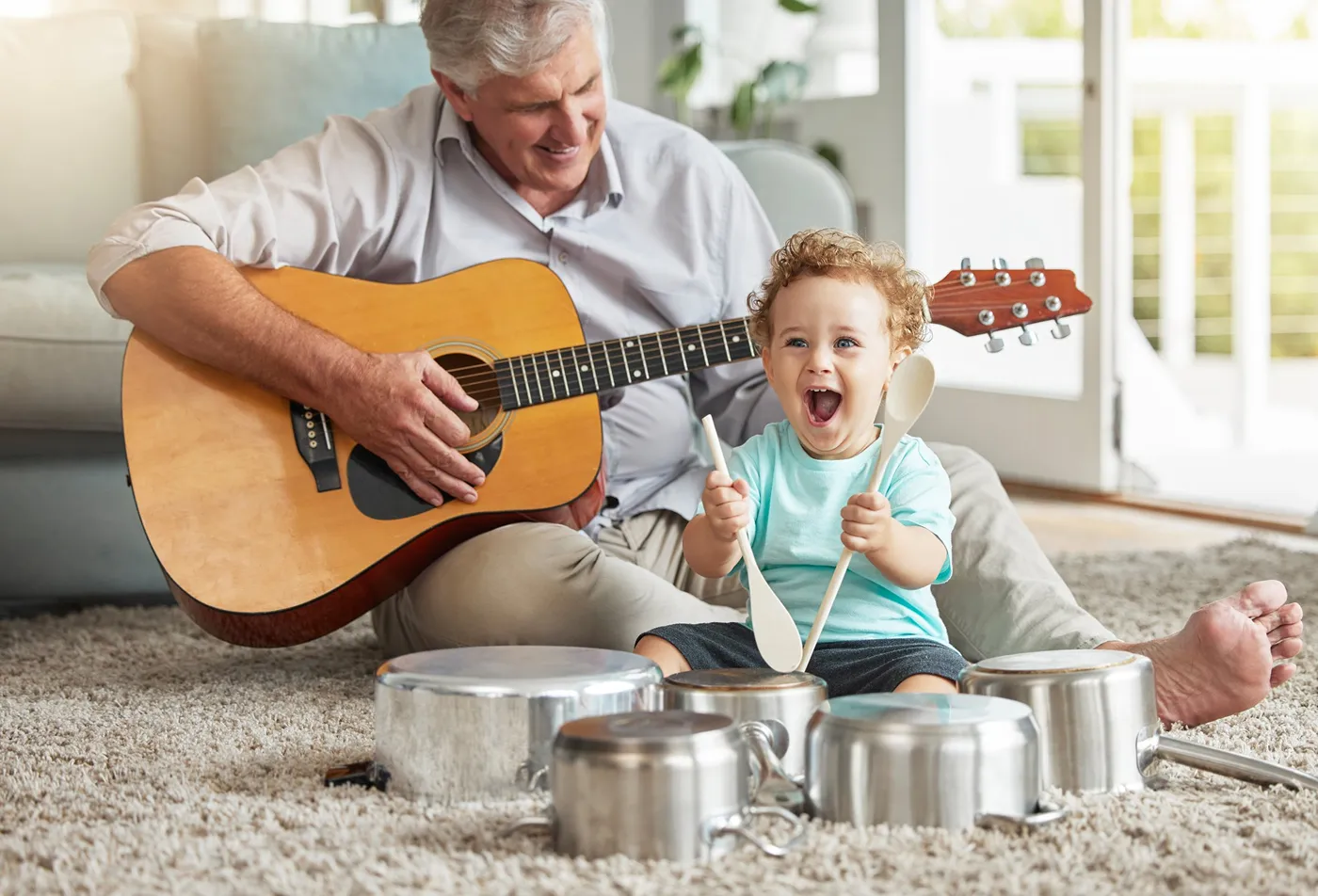 Smiling older man plays guitar while excited toddler uses spoons on pots as drums indoors.