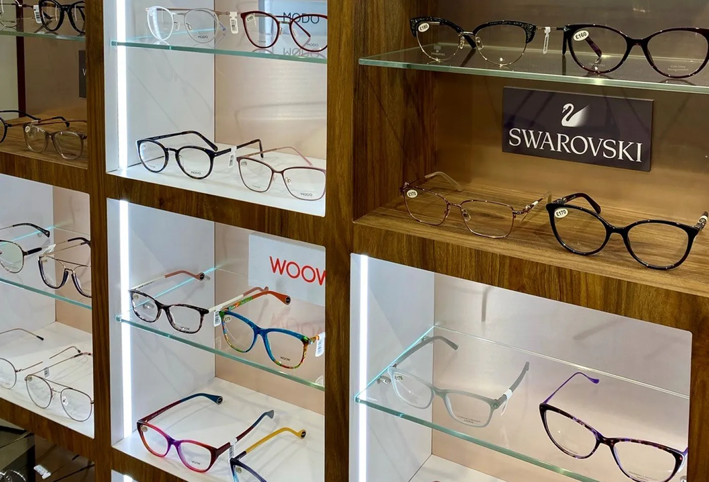 Eyeglasses frames from brands including Swarovski and Woow displayed on lit shelves