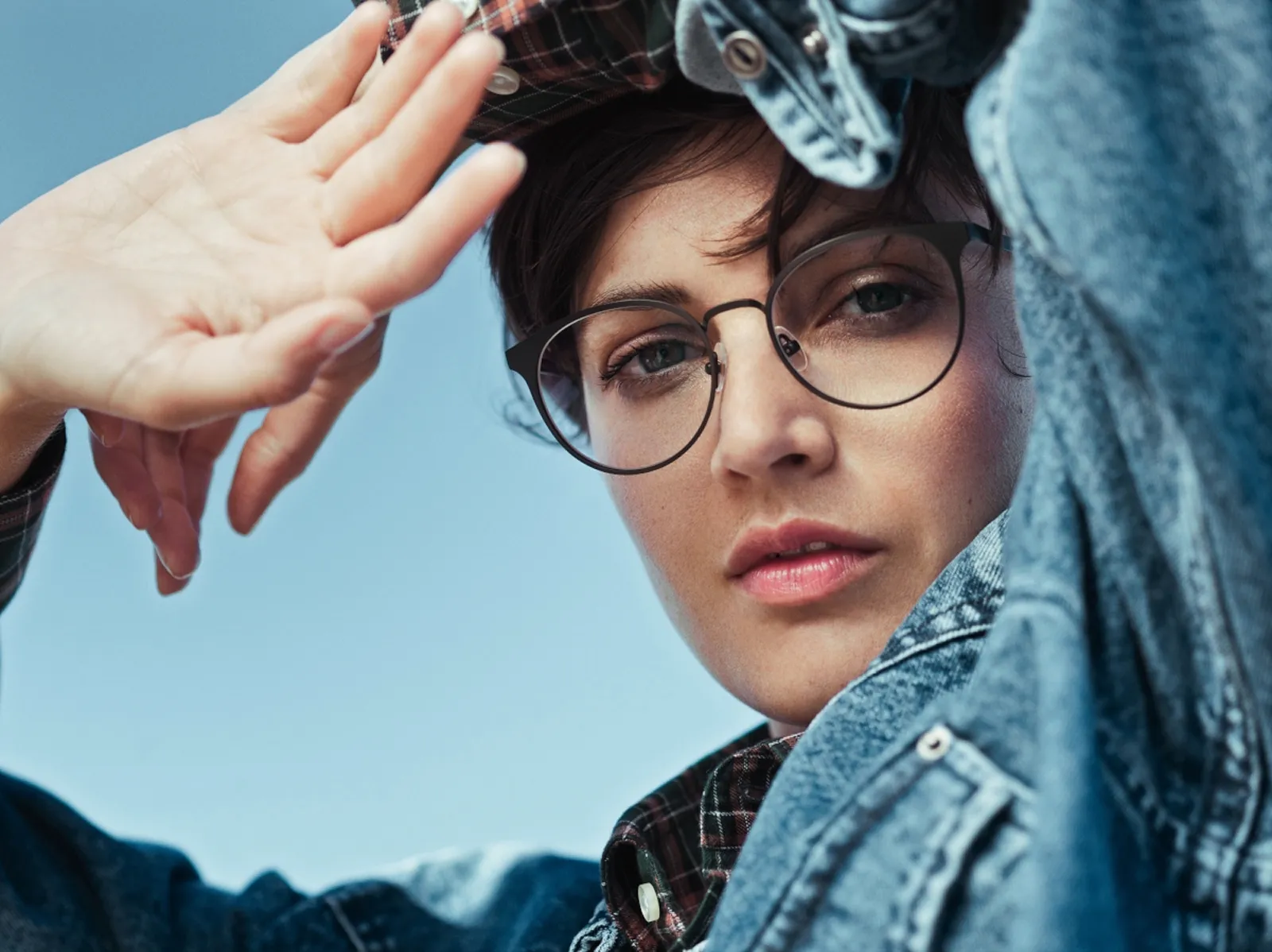 Close-up of a person wearing glasses and a denim jacket, raising a hand against a blue sky.
