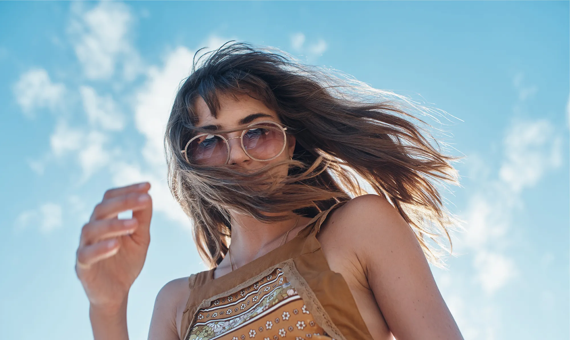 Woman wearing sunglasses with hair blowing in wind against a blue sky with scattered clouds.