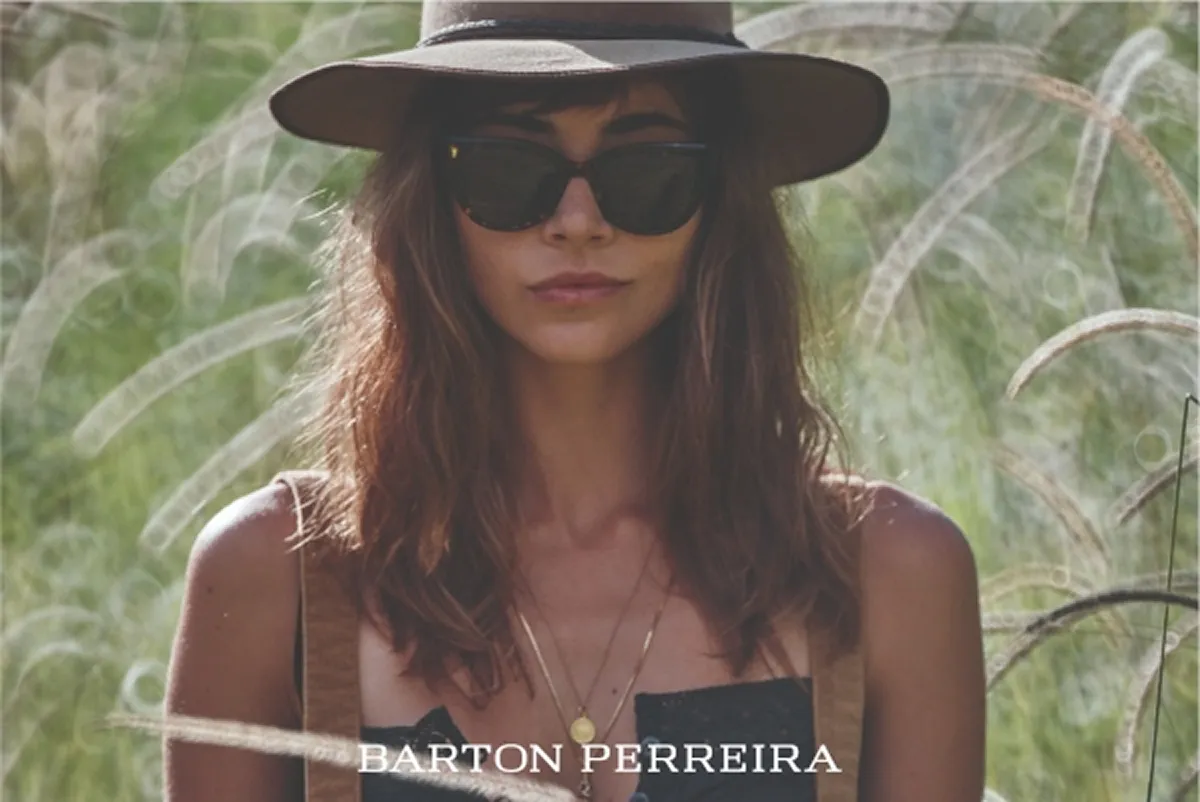 Woman wearing a wide-brimmed hat and dark sunglasses standing in tall grass outdoors.