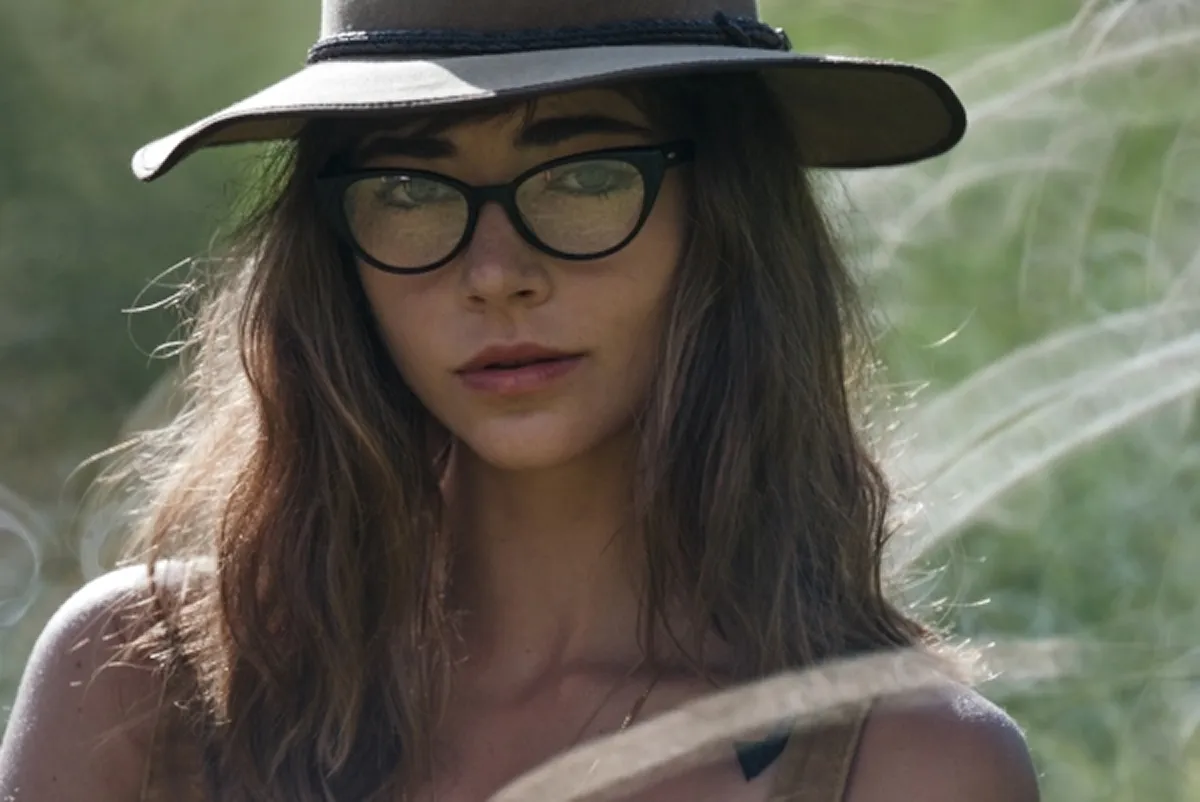 Young woman with long brown hair wearing black glasses and a wide-brimmed hat outdoors.