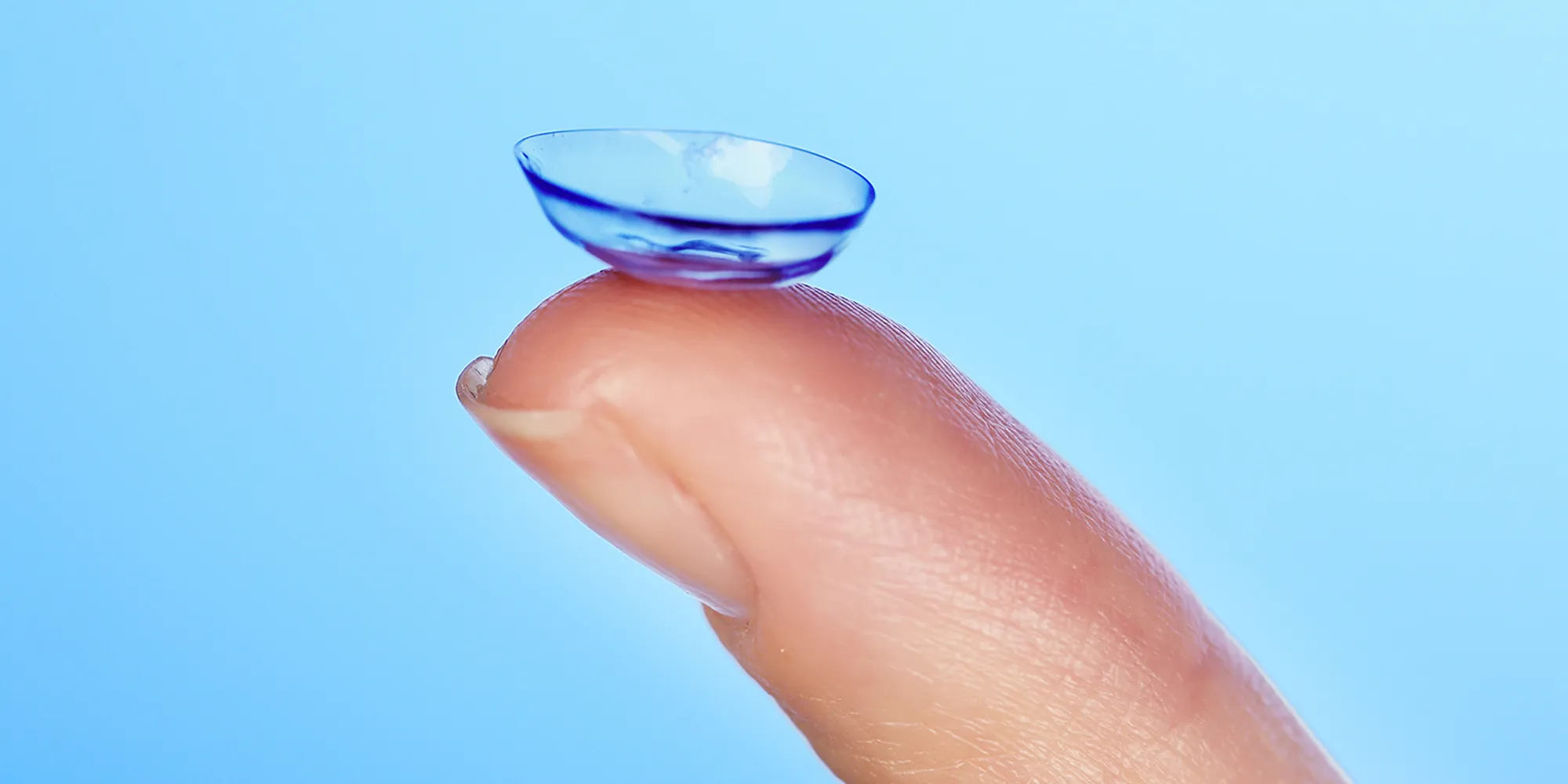 Close-up of a contact lens balanced on the tip of a finger against a blue background.