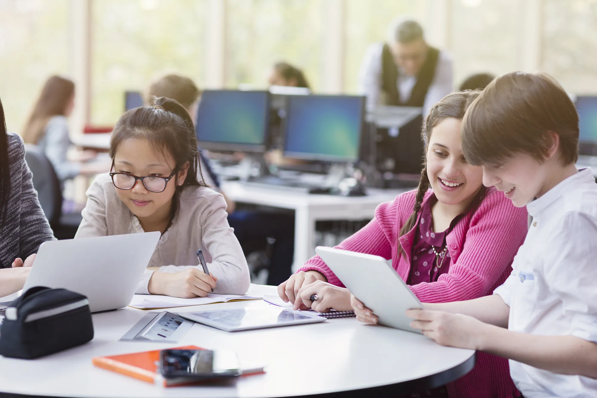 Three students working at a round table with laptops and tablets in a computer lab.