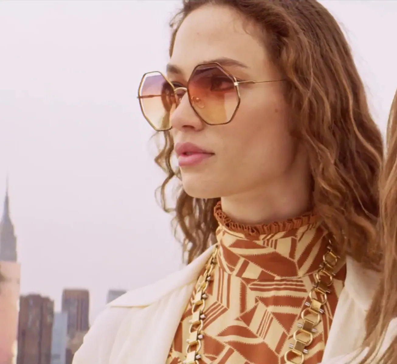 Woman wearing large hexagonal sunglasses, patterned orange top, and gold chain necklace with cityscape background.