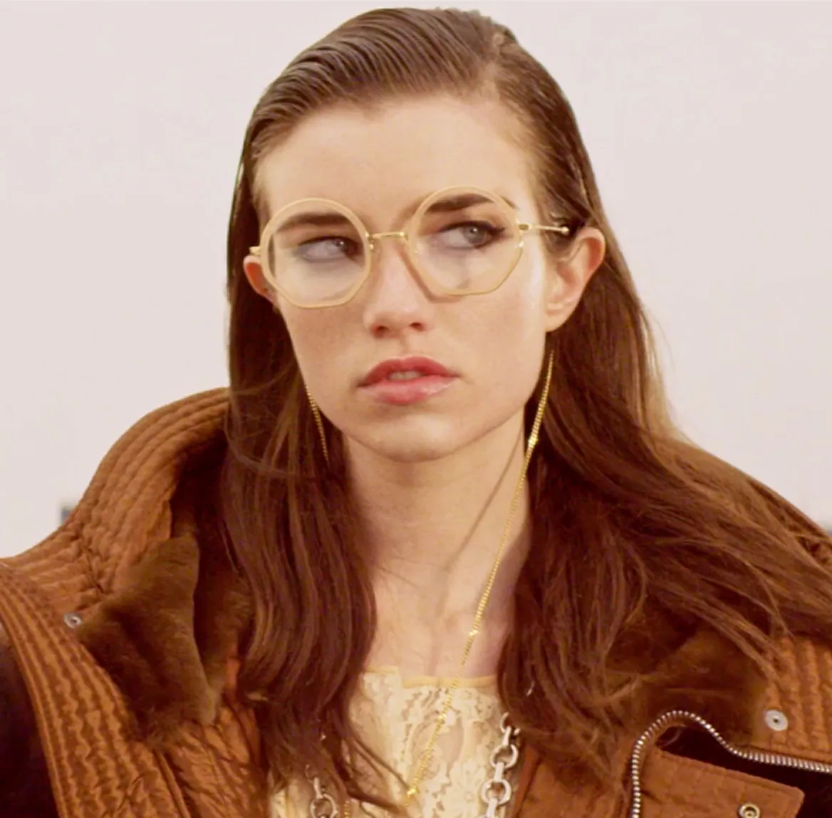 Woman with long brown hair wearing large octagonal glasses and a brown textured jacket.