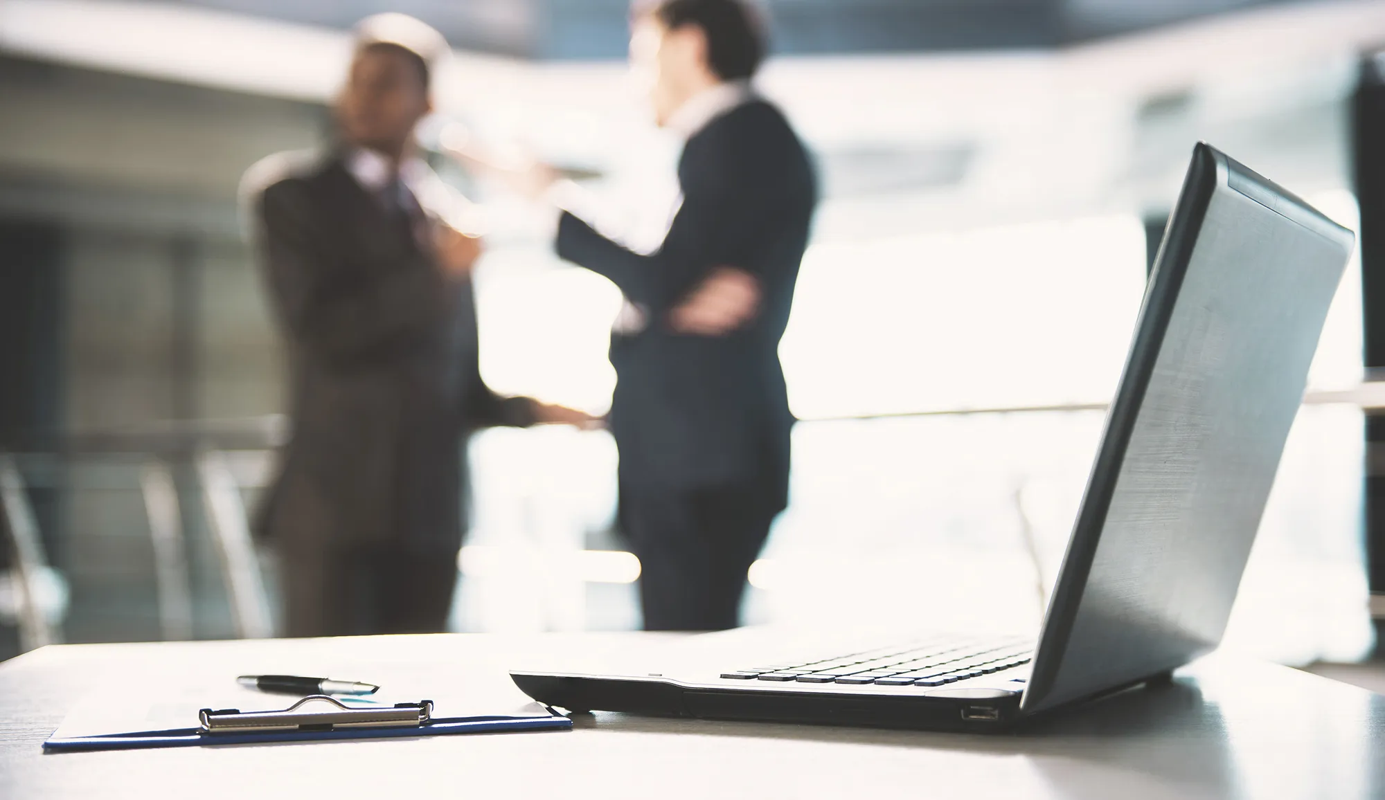 Open laptop on a desk with clipboard and pen, two people in business suits talking blurred in background.