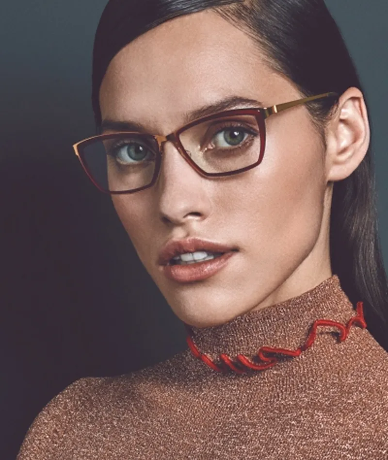 Close-up portrait of a woman with green eyes wearing brown rectangular eyeglasses and a brown glittery top with red trim.