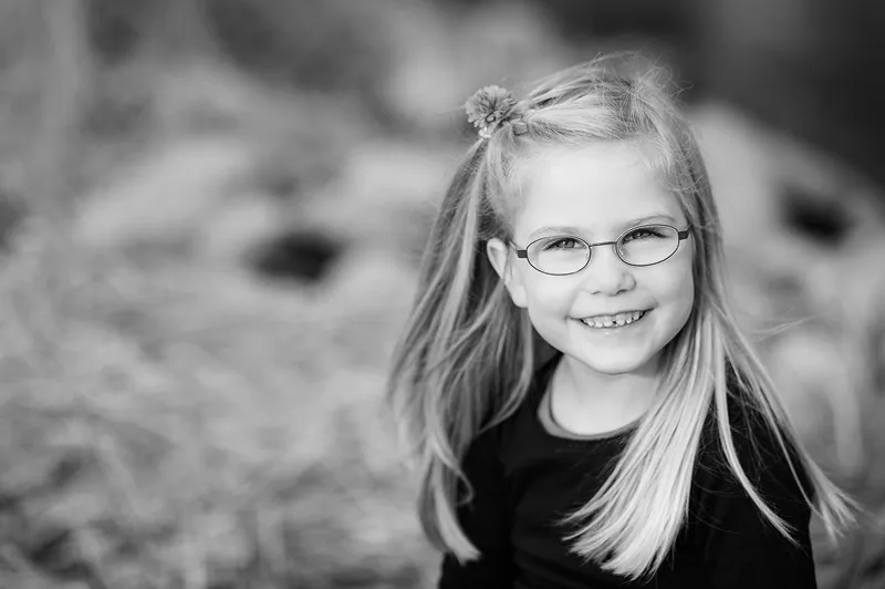 Smiling young girl with straight hair, glasses, and a flower hair clip wearing a black top.