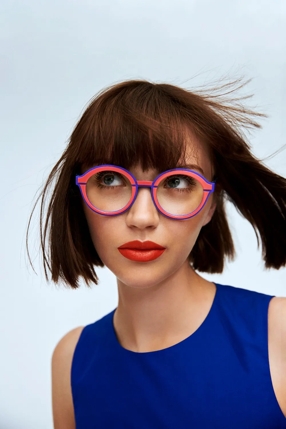 Young woman with short brown hair wearing round red and blue glasses and bright red lipstick in blue top.