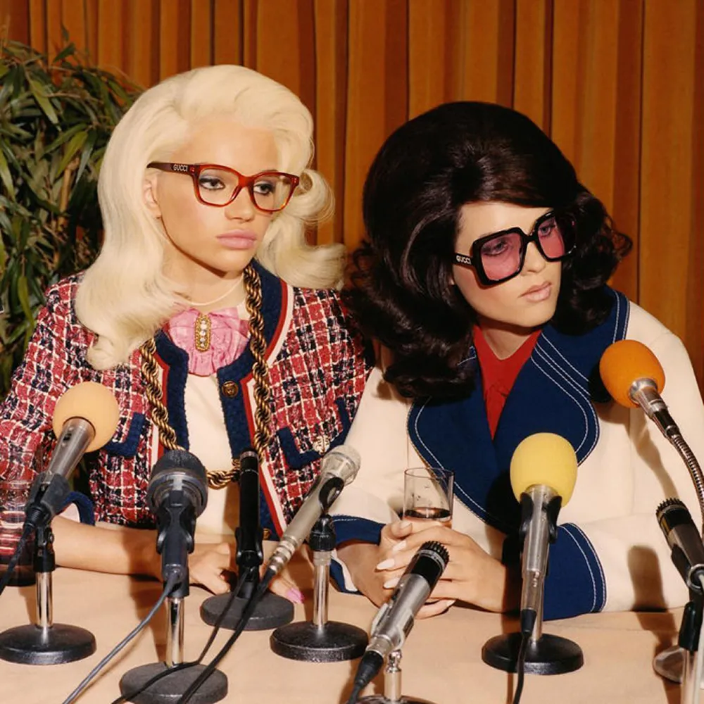 Two women with vintage hairstyles and Gucci glasses sit at a table with microphones, one holding a glass of water.