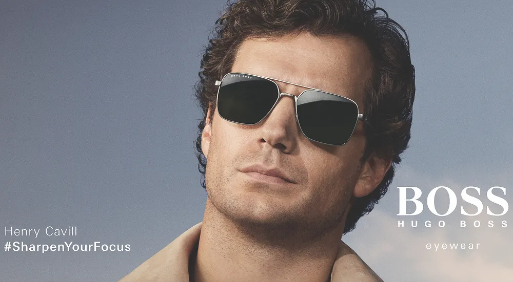 Henry Cavill wearing black Hugo Boss sunglasses with beige coat and a blue sky background.