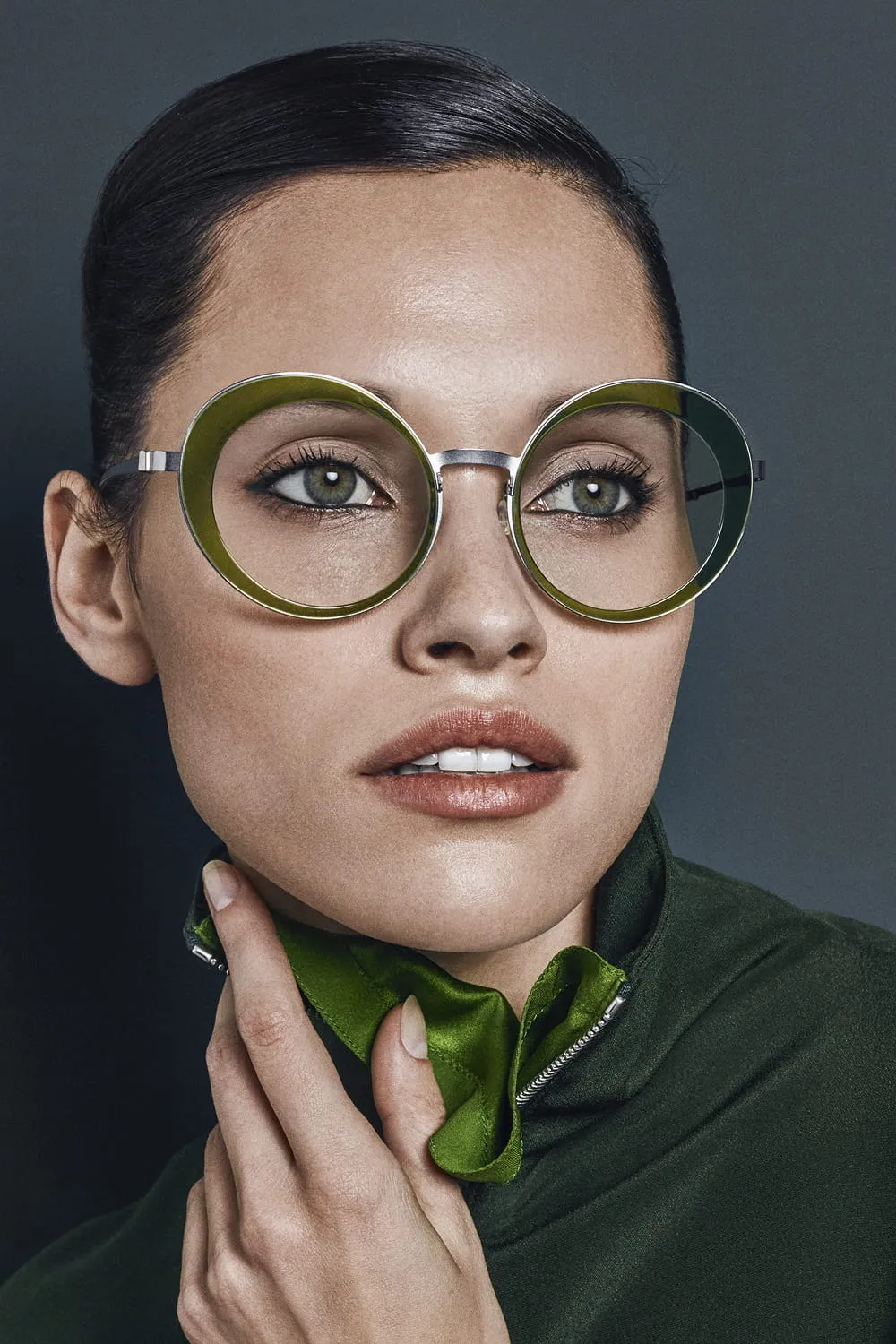 Close-up of a woman wearing large round greenish eyeglasses and a dark green outfit.