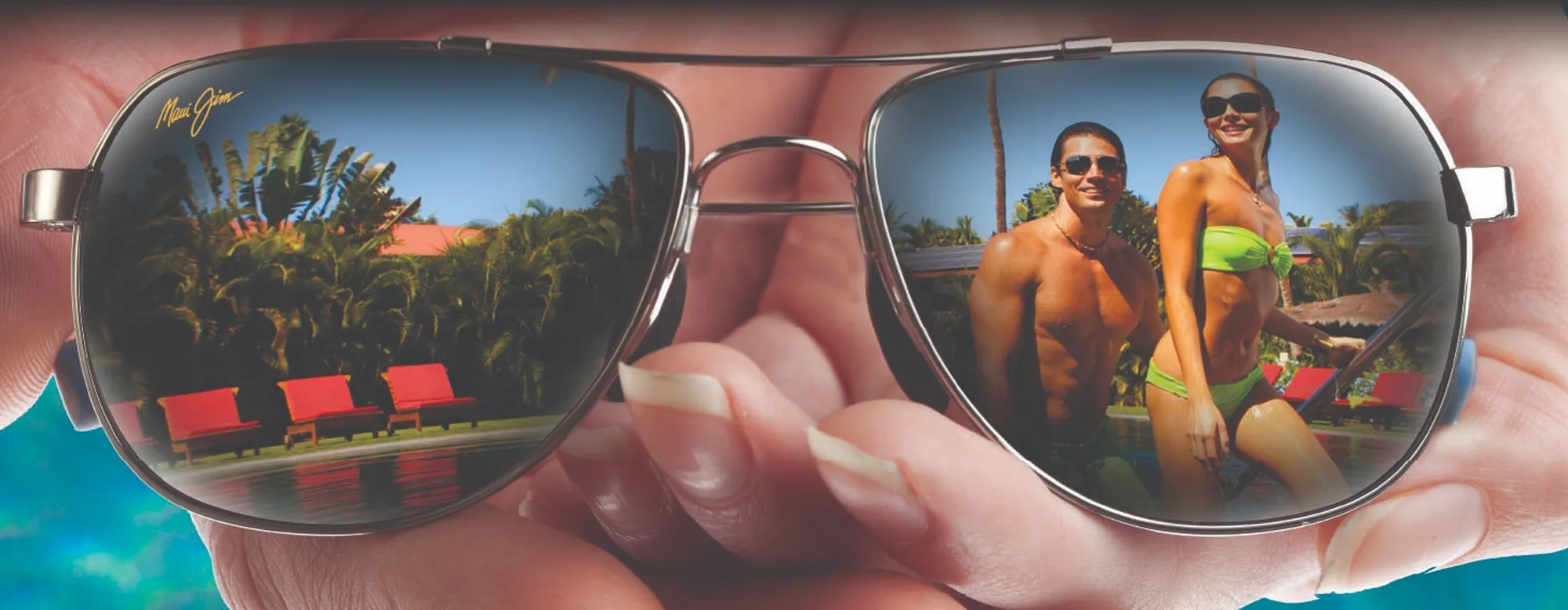 Reflection in sunglasses of a smiling man and woman by a pool with red lounge chairs and palm trees.