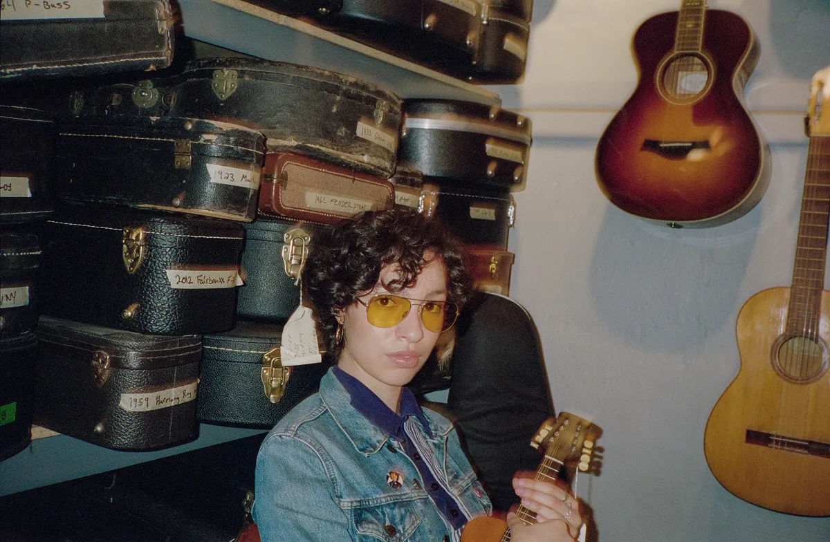 Person with curly hair and yellow glasses holding a small string instrument surrounded by guitar cases and hanging guitars.
