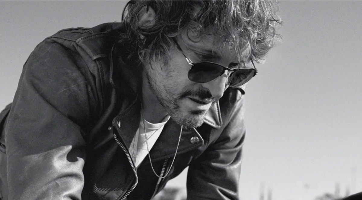 Black and white photo of a man wearing sunglasses and a leather jacket looking down.