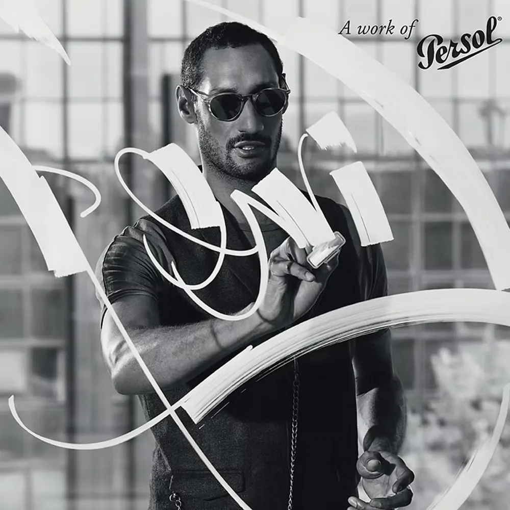 Man wearing sunglasses painting abstract white brush strokes on glass in black and white photo.
