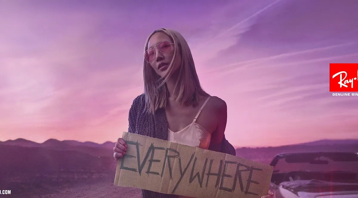 Woman wearing pink sunglasses holding a cardboard sign that says 'EVERYWHERE' with a mountainous sunset background.