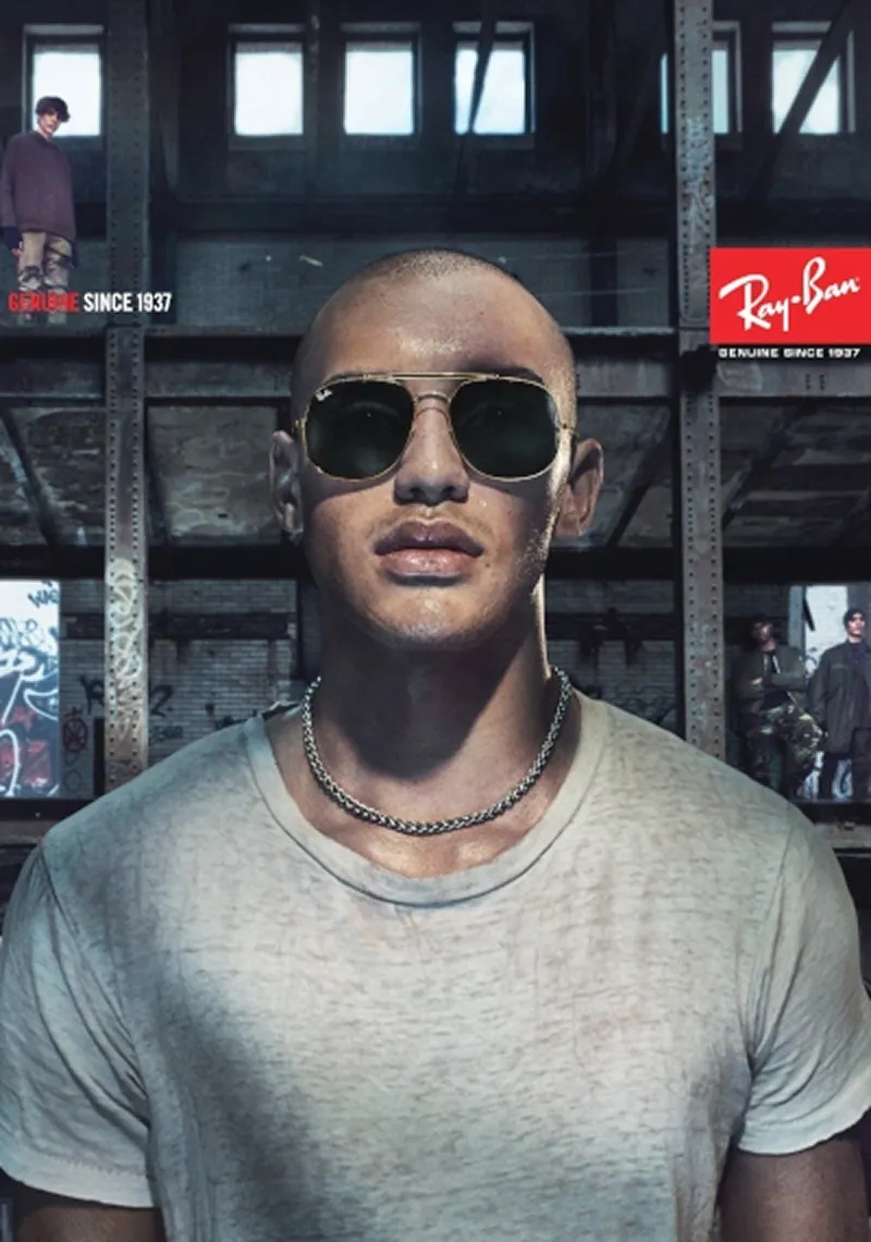 Man wearing gold-framed Ray-Ban sunglasses and a chain necklace in an industrial setting.