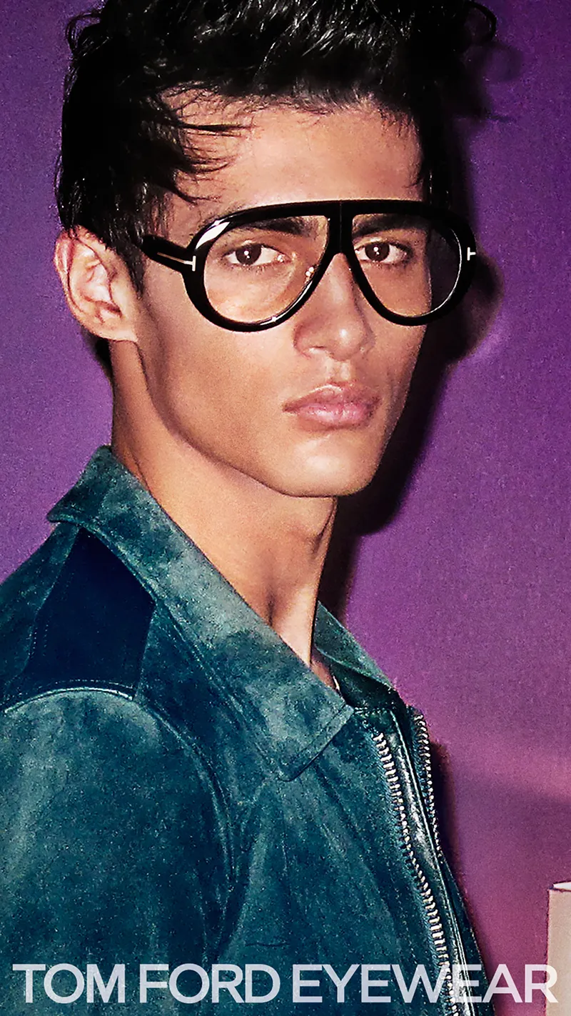 Young man wearing large black Tom Ford eyeglasses and a dark green jacket against a purple background.