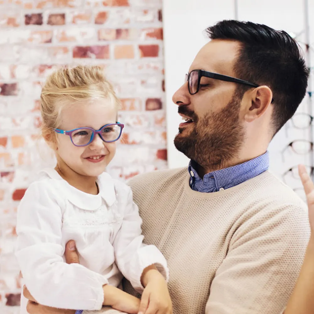 Man holding smiling young girl wearing blue glasses in front of a brick wall.