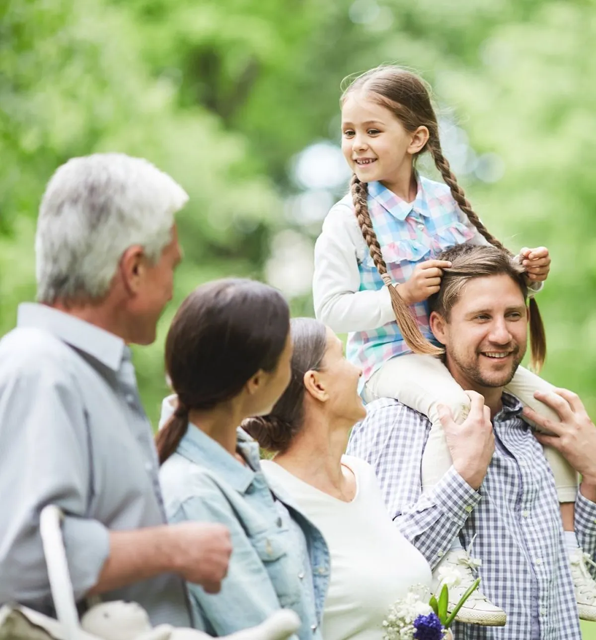 Smiling multi-generation family outdoors with a young girl sitting on a man's shoulders in a green park.