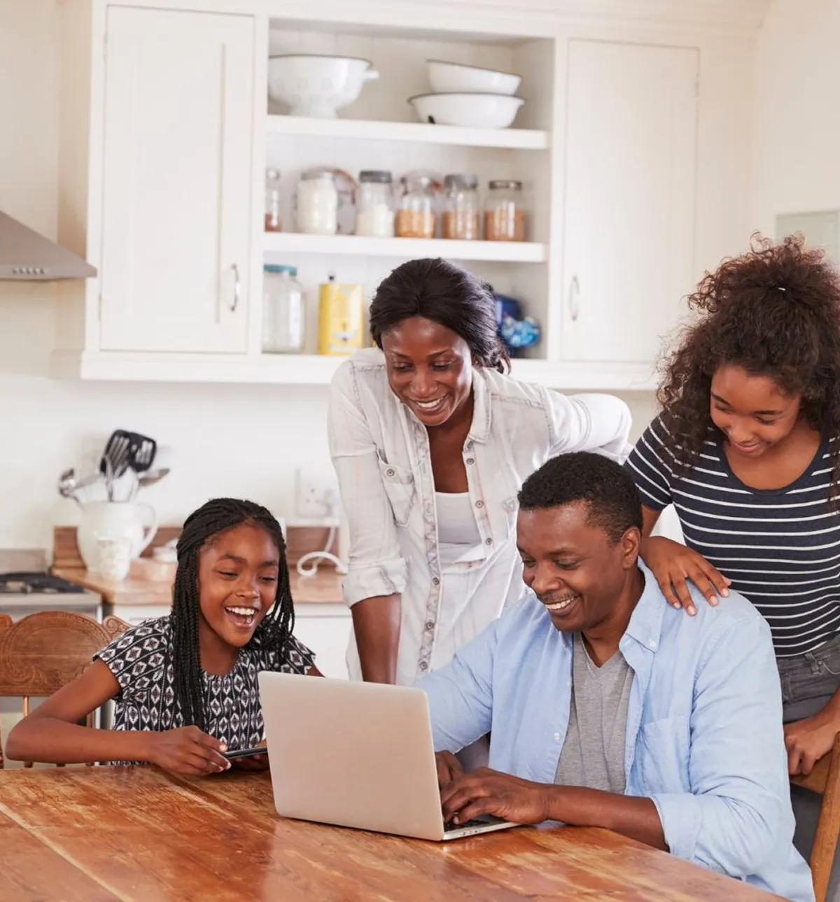 Smiling family of four gathered around a laptop in a kitchen, enjoying time together.
