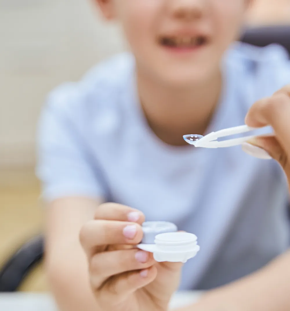Child holding a contact lens case while a hand uses tweezers to hold a contact lens.