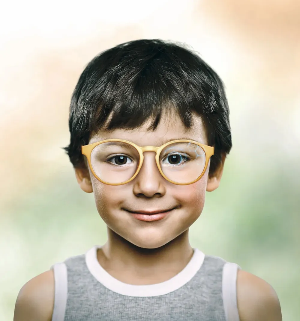 Smiling young boy with short dark hair wearing round yellow glasses and a gray sleeveless shirt.