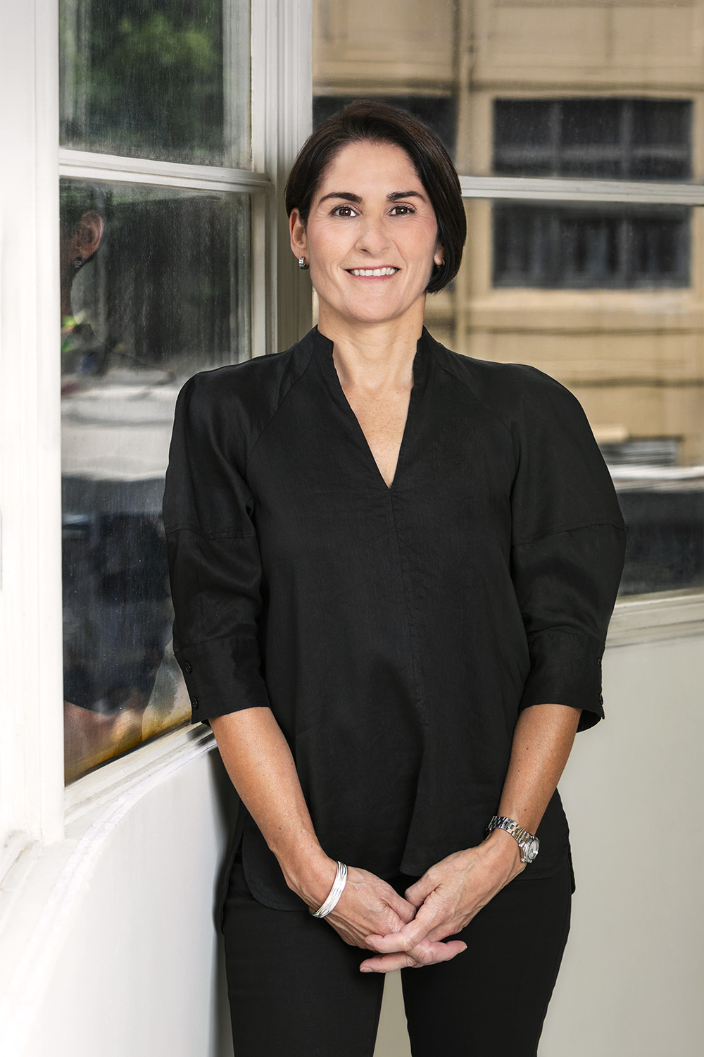 Woman with short dark hair wearing a black top, standing by a window with hands clasped.
