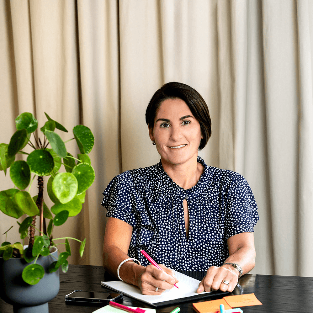Woman in navy blue blouse with white stars writing in notebook at a dark wood desk with a green plant.