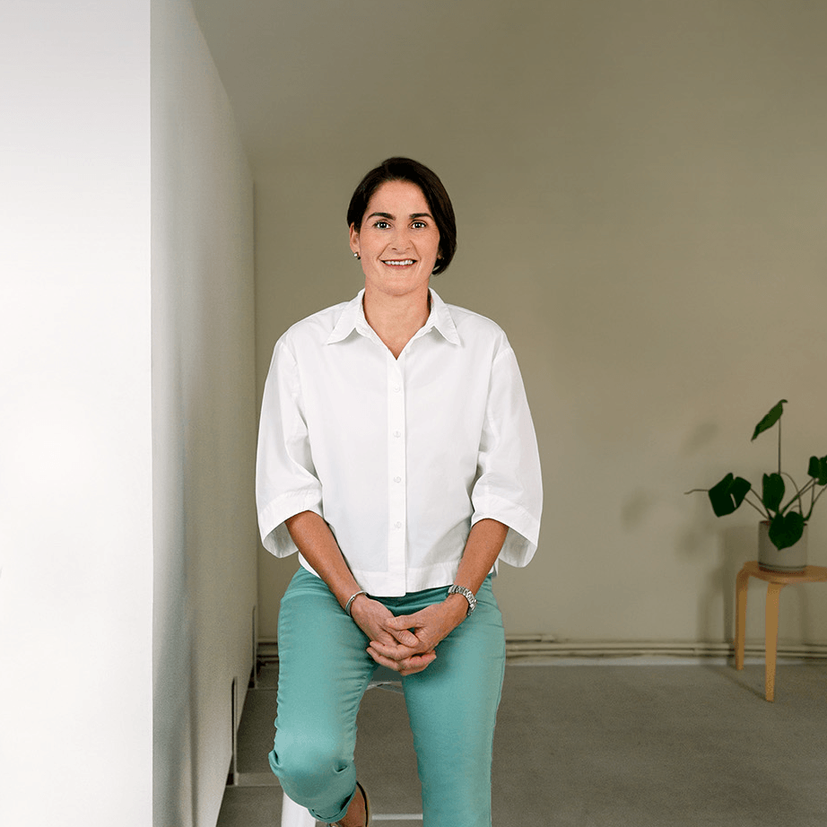Woman with short dark hair wearing a white shirt and teal pants sitting on a stool against a light wall.