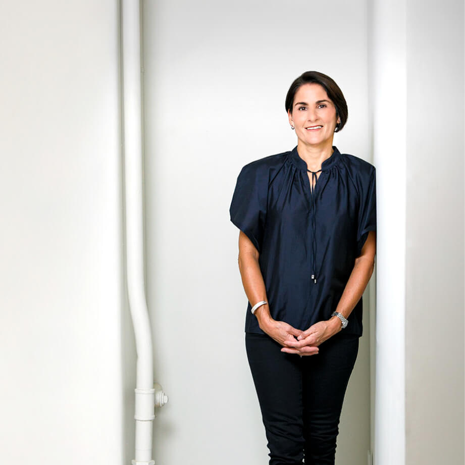 Smiling woman in black blouse and pants standing against a white wall near a vertical pipe.